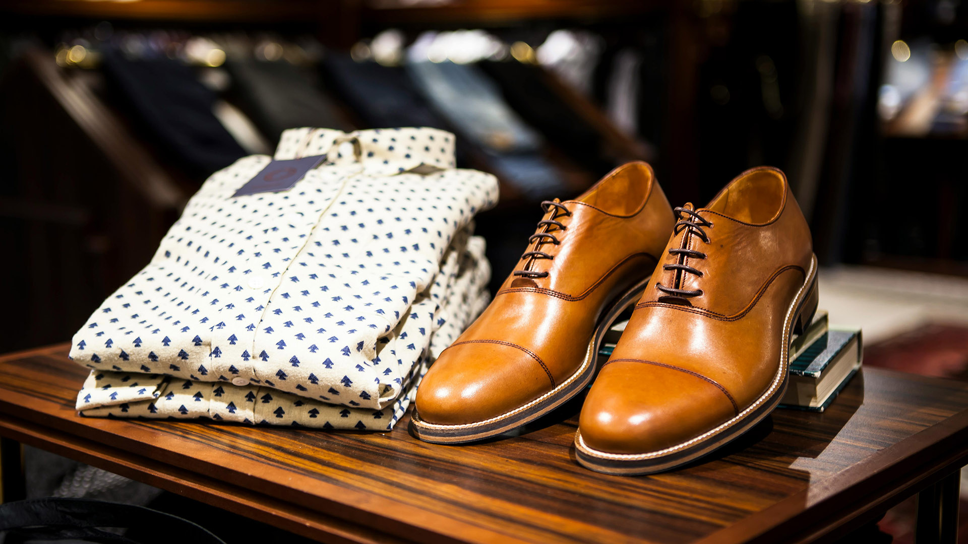 Braune Lederschuhe neben gefalteten Hemden mit blauem Muster, auf Holztisch in elegantem Geschäft.
