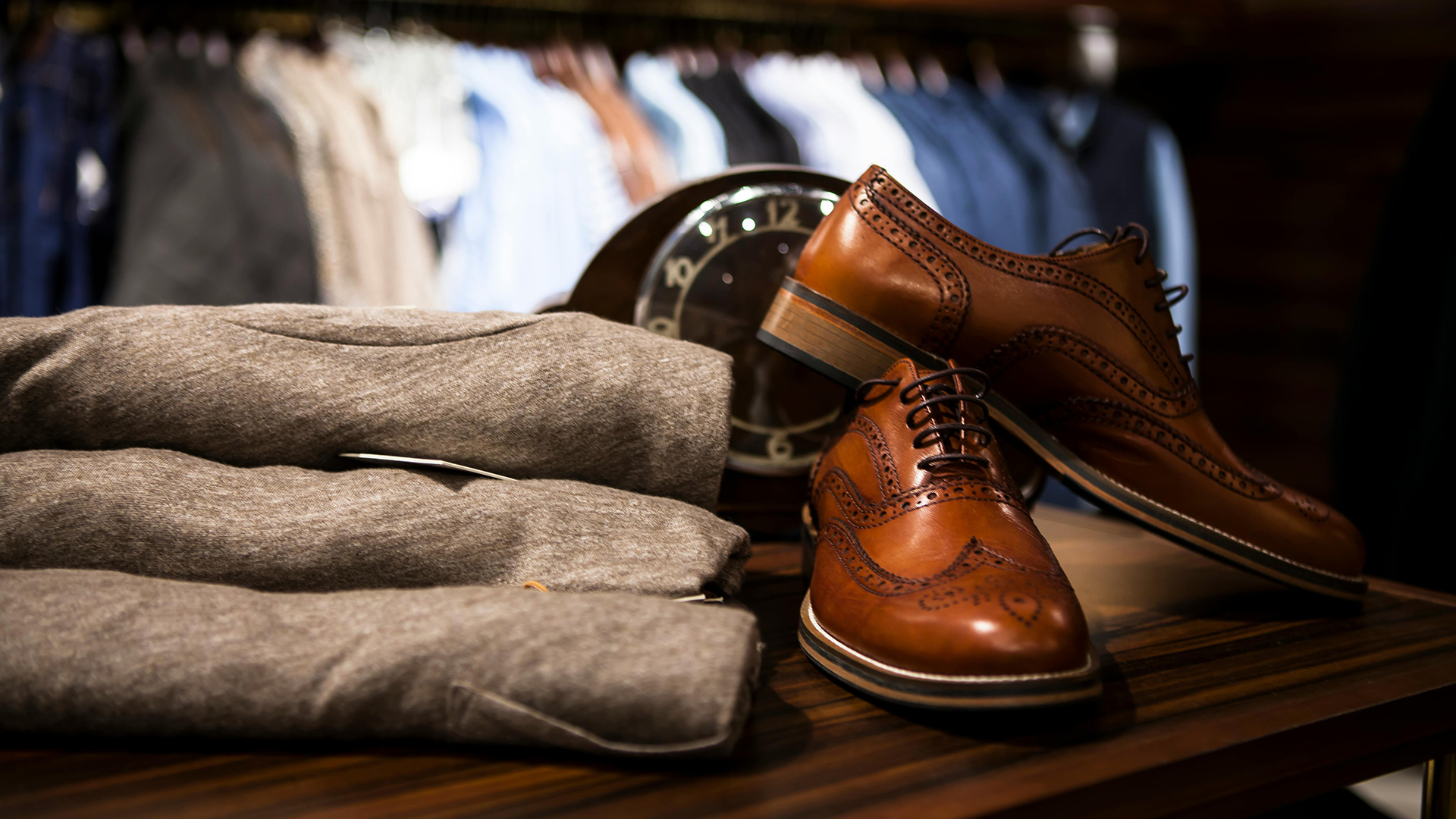 Braune Schuhe und graue Kleidung auf Holztisch vor unscharfem Hintergrund mit Uhr und Kleidern.